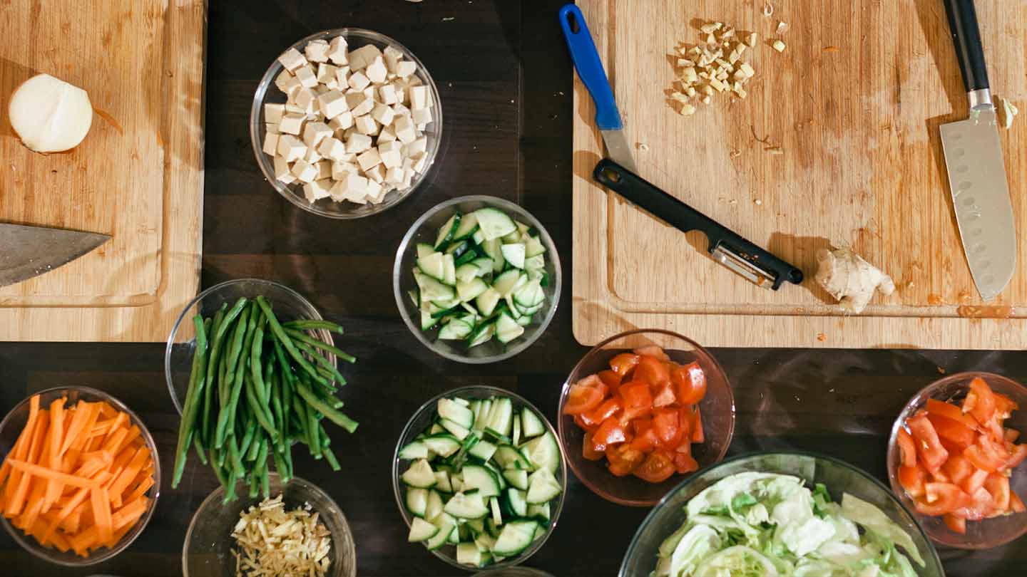 Mesa con tablas de cortar, tazas con alimentos cortados de distintas formas, alrededor, manos de mujeres