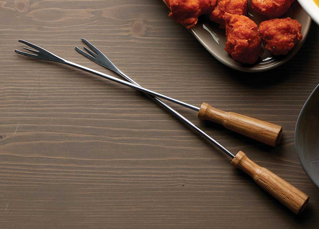 Sobre una mesa dos tenedores para fondue, salsa de queso, Nuggets de pollo y papas artesanales