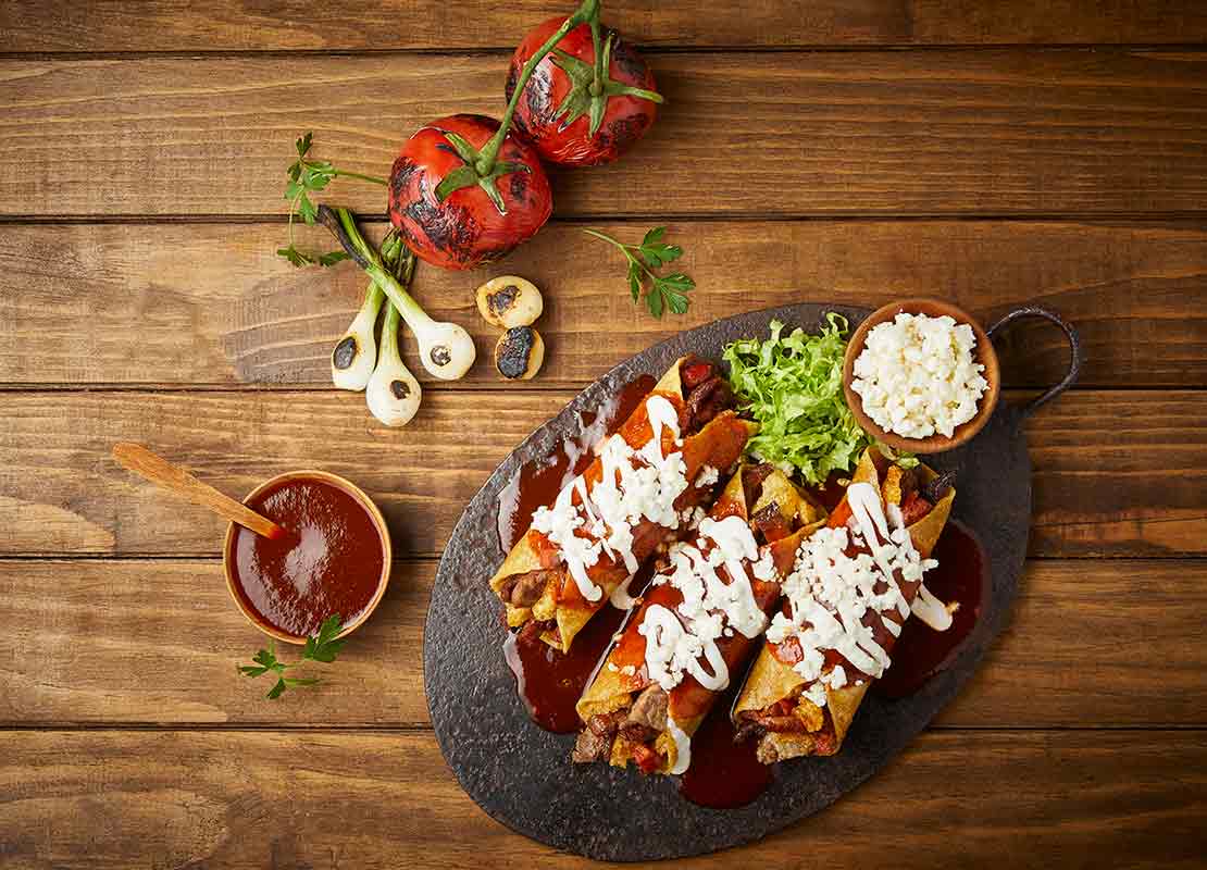 Tres burritos de carne en una placa de piedra negra natural, al lado cuenco con salsa, jitomates y ajos