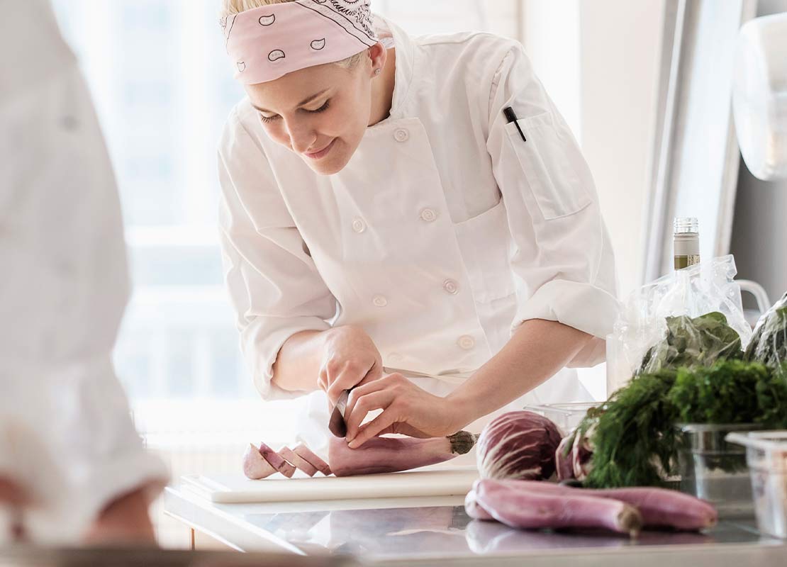 Una chef corta vegetales cuidadosamente al fondo de una cocina, al lado utensilios de acero inoxidable