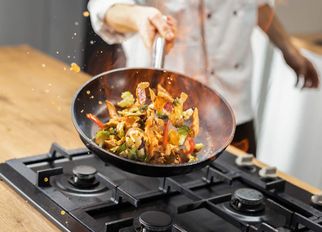 A domicilio, un chef saltea vegetales en una sartén cuya flama se proyecta sobre la comida