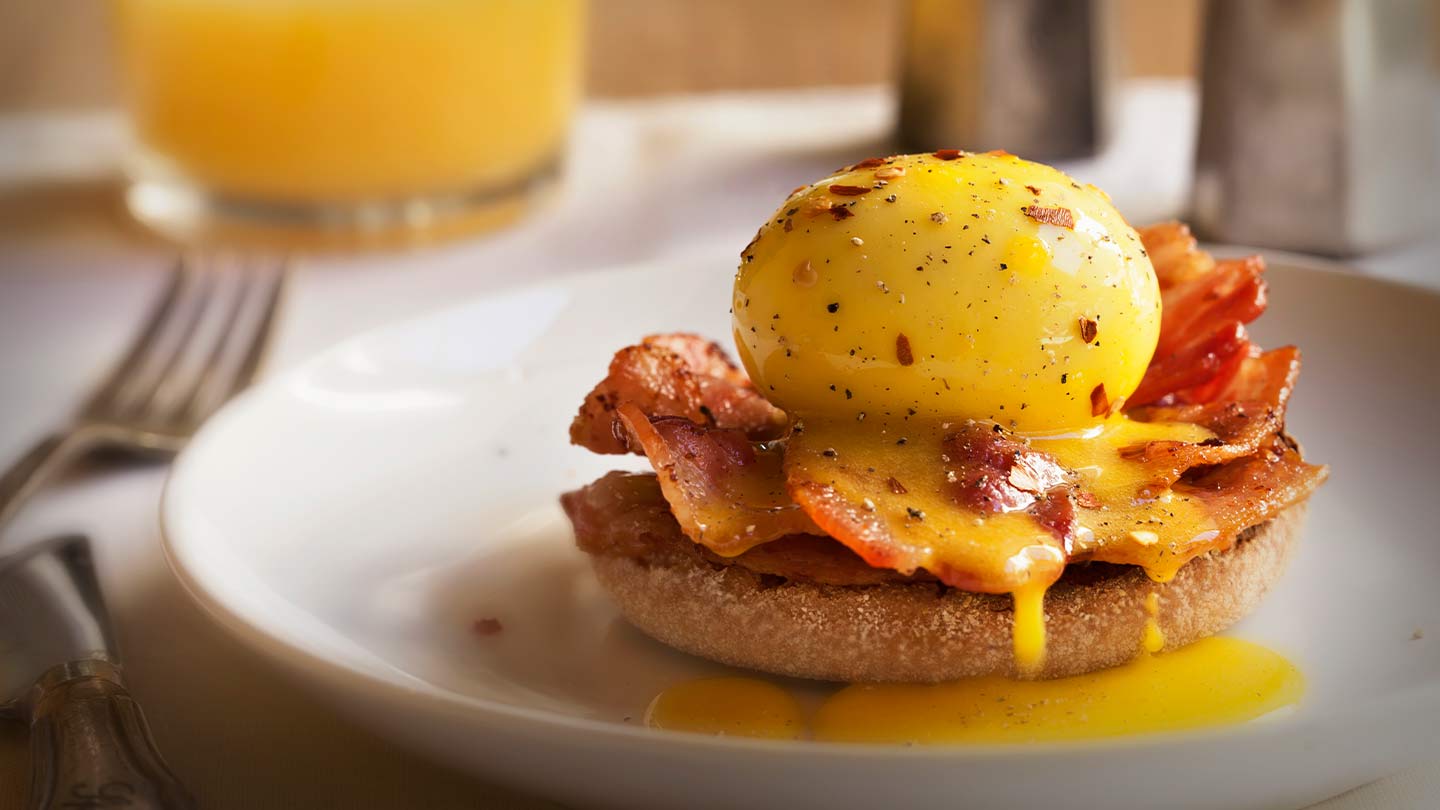 Huevo benedictino con bacon y salsa holandesa sobre un muffin inglés, al fondo jugo y saleros
