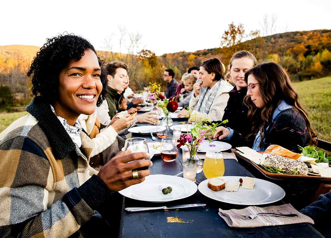 Grupo de amigos comen al aire libre sentados en una mesa de madera, rectangular y larga