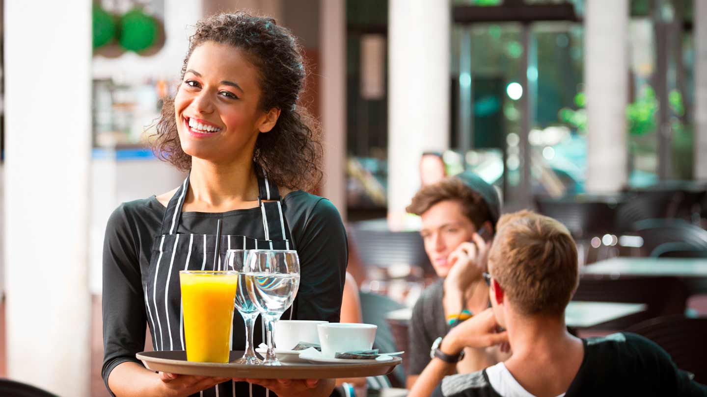 Mesera sonríe mientras sostiene una bandeja con copas, tazas y jugo, al fondo comensales sentados