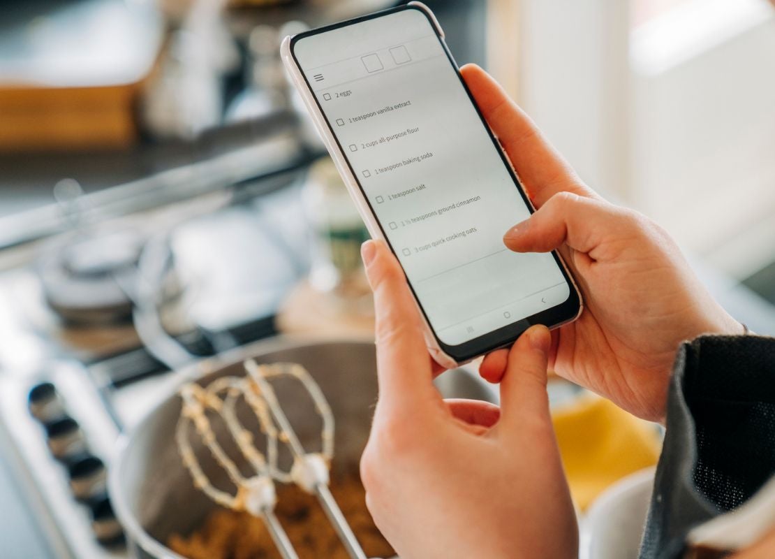 Manos de mujer sostienen un celular que muestra una lista de ingrediente, al fondo utensilios de cocina