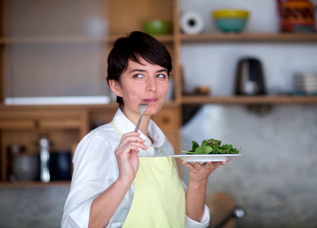 En una cocina, una chef sostiene un tenedor en su boca mientras en la otra mano tiene un plato de ensalada