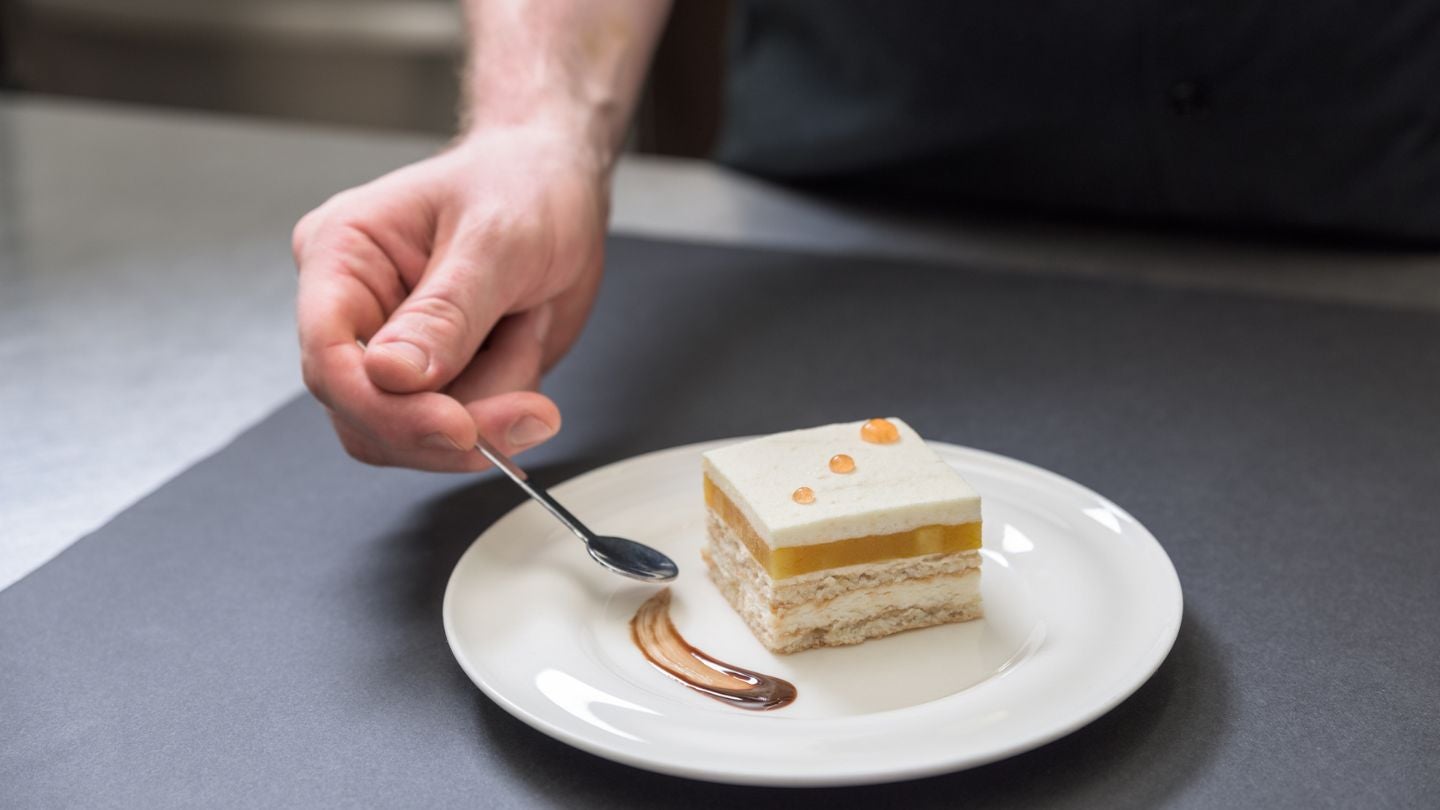 Mano de chef pastelero sostiene una cuchara con la que decora el plato de un postre con salsa