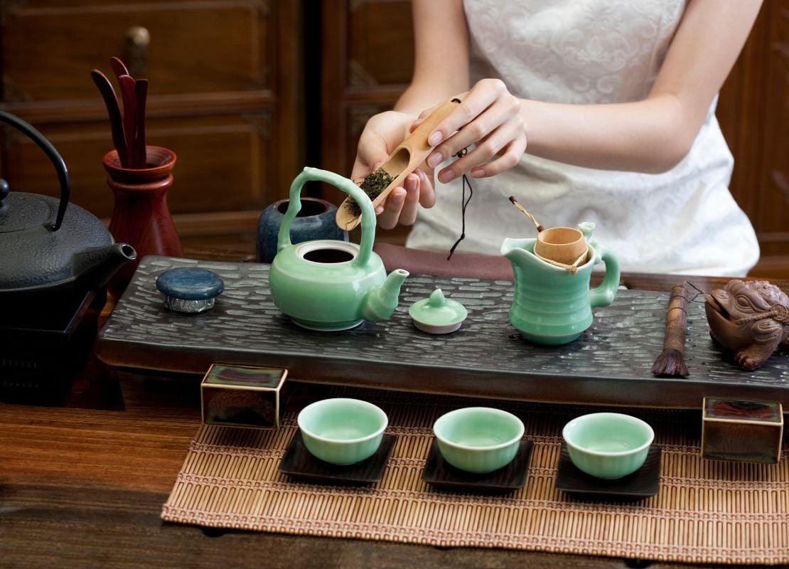 Mujer vierte hojas en una tetera de porcelana sobre una mesa y rodeada de otros utensilios para elaborar té