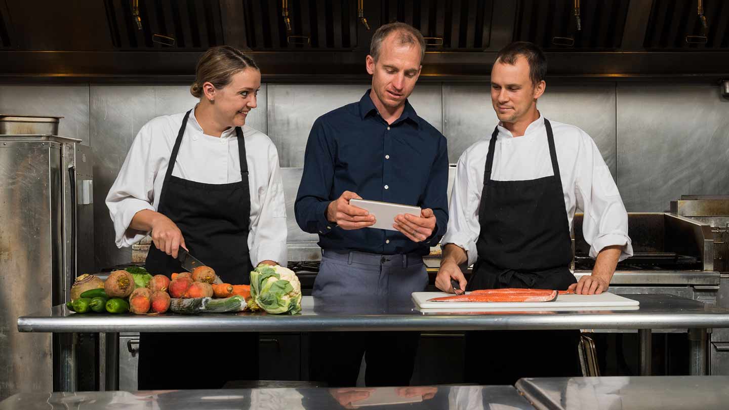 Dos chefs y un gerente de cocina revisan una tablet, rodeados de ingredientes frescos en una cocina profesional