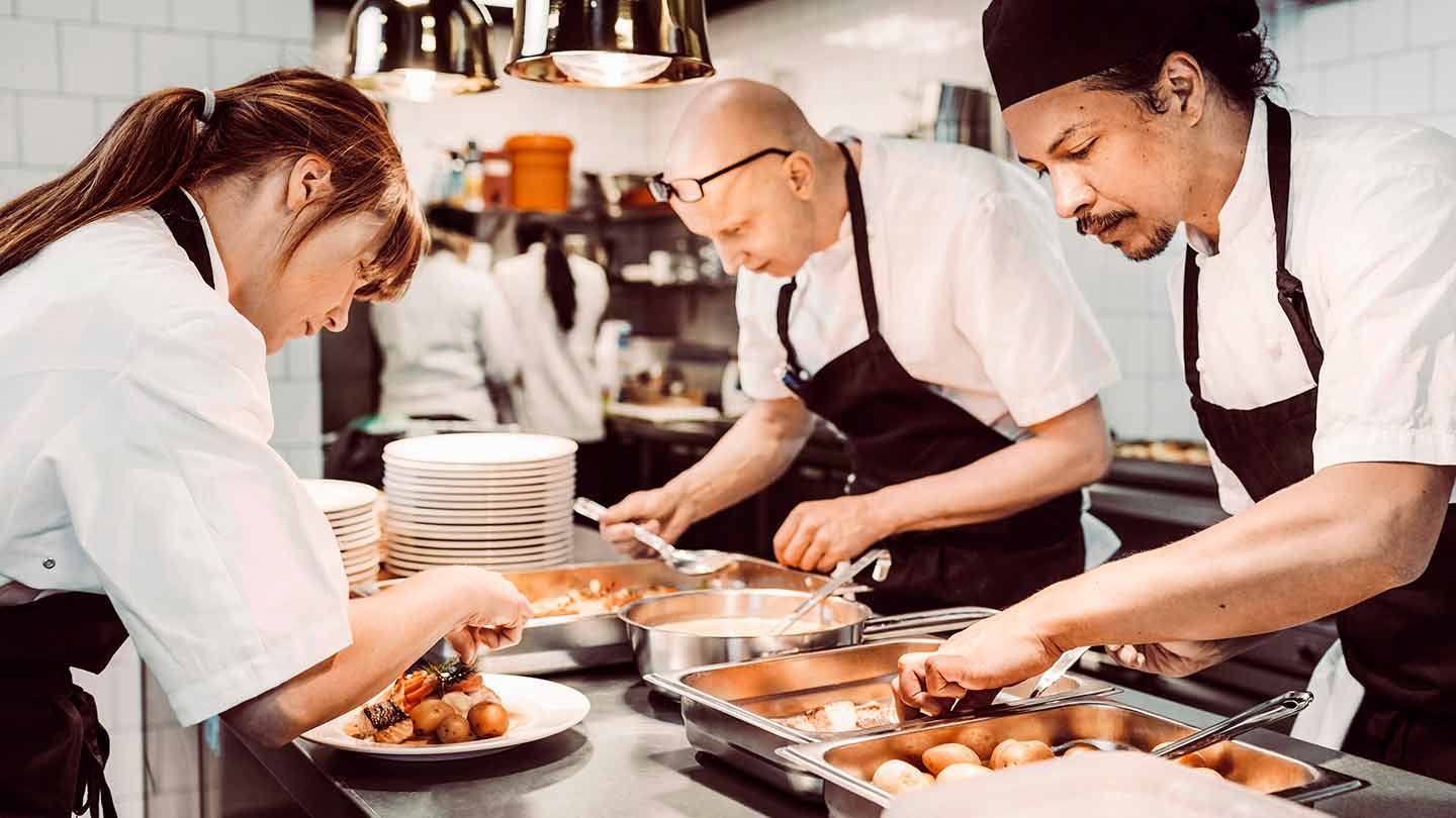 3 chefs en una cocina comercial seleccionan alimentos de la mesa para emplatar las preparaciones de sus clientes