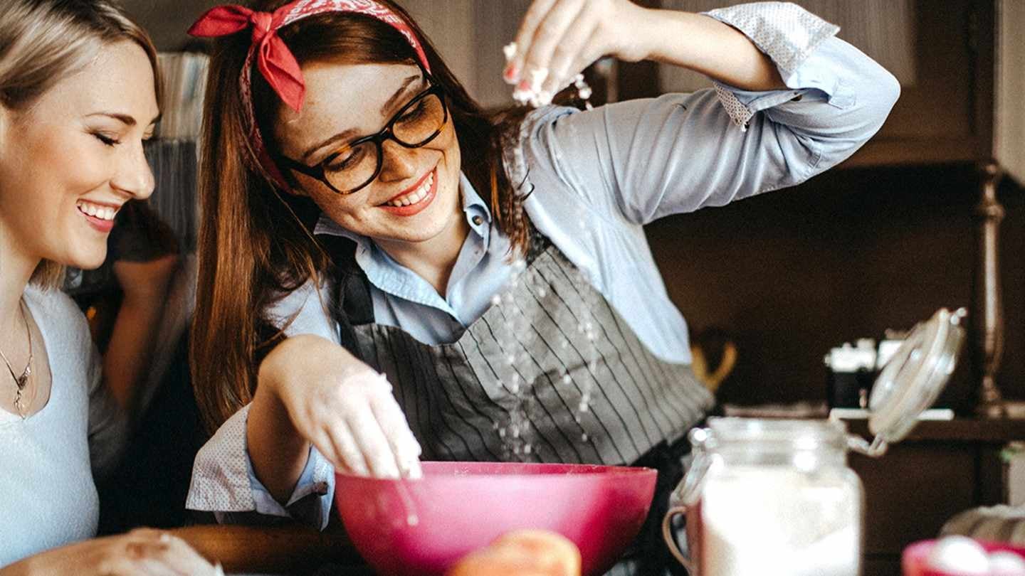 Repostera espolvorea harina en un bowl sobre el mesón de una cocina, rodeado de otros ingredientes