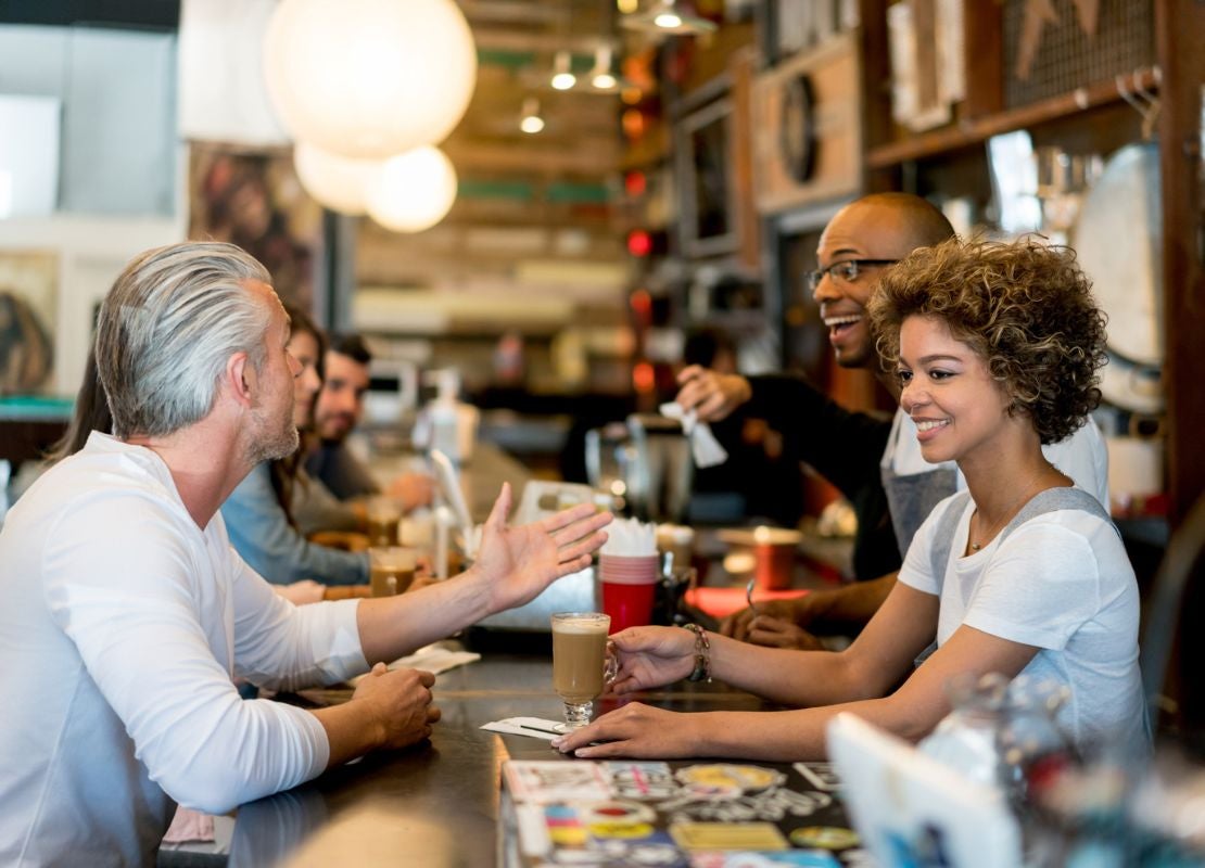 Cliente recibe una bebida de café e interactúa con dos meseros sobre la barra de la cafetería