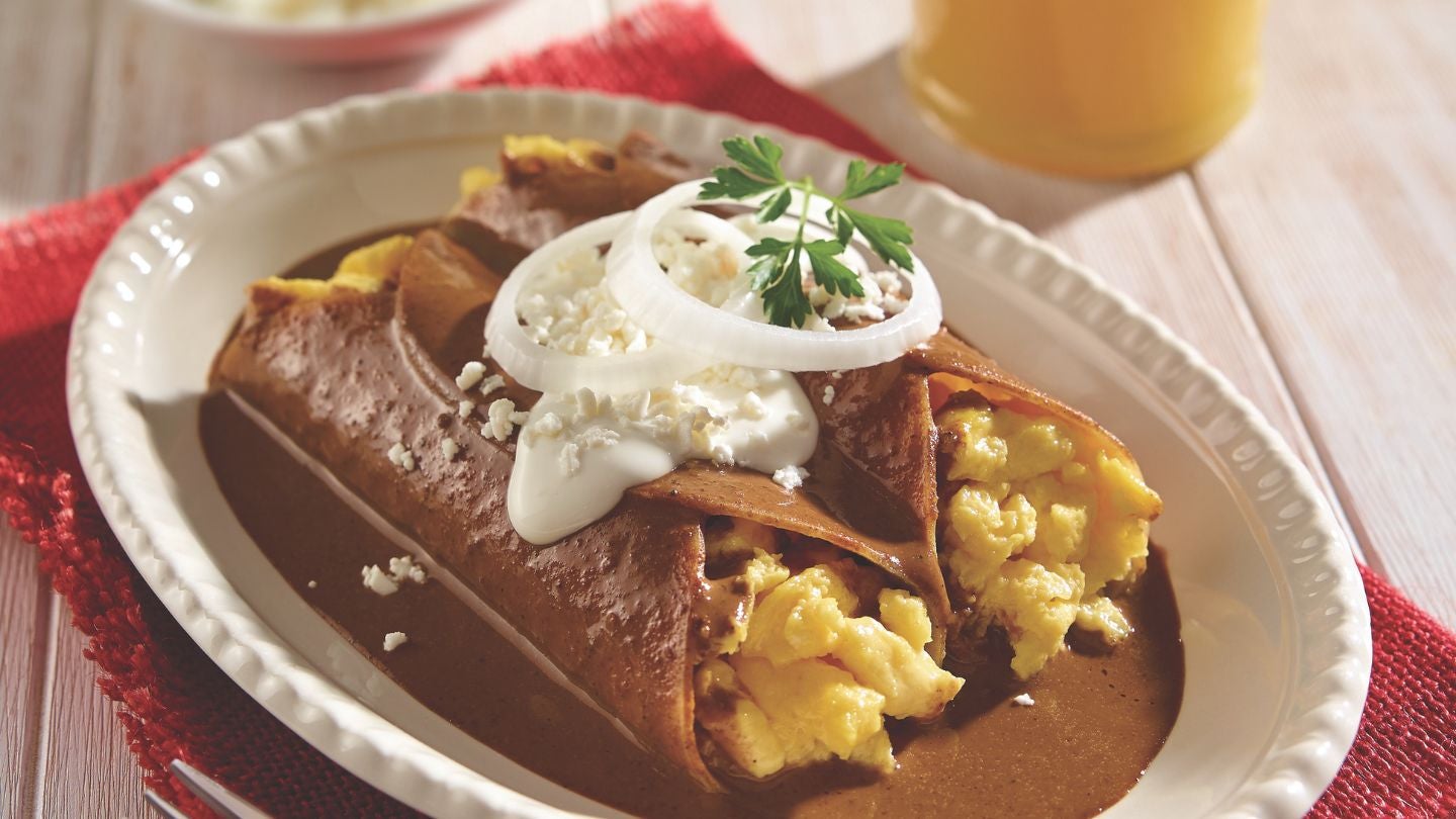 Enmoladas de huevo cubiertas con mole y crema, junto a un vaso de jugo.