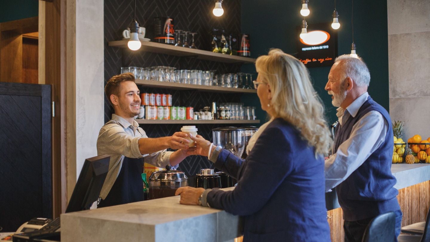 Barista entrega una bebida a una clienta en cafetería mientras otro hombre espera