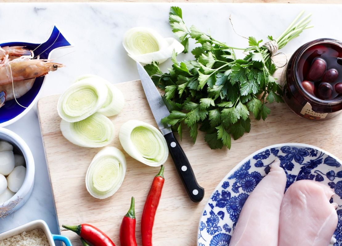 Ingredientes frescos como pollo, verduras y camarones junto a un cuchillo sobre una tabla