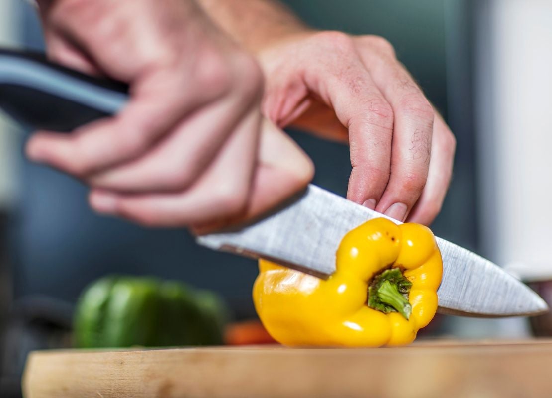 Manos cortan un pimentón amarillo con un cuchillo para verduras sobre una tabla.