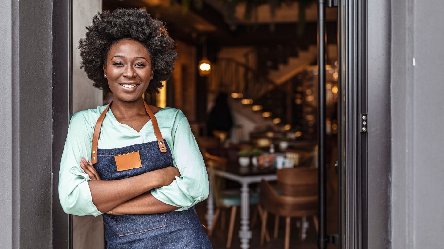 Dueña de negocio gastronómico con delantal frente a la puerta de su establecimiento