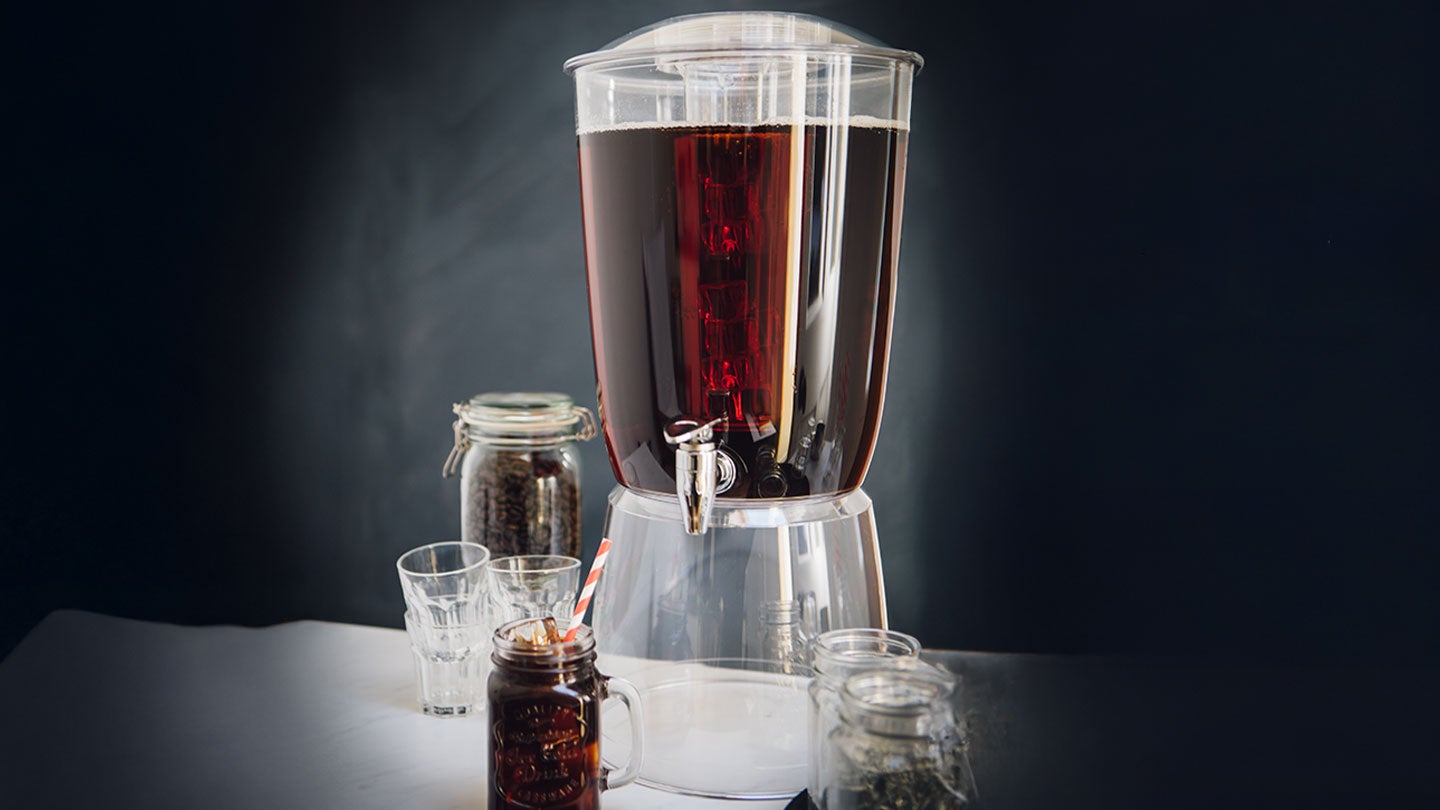Dispensador de café cold brew junto a vasos y frascos con granos de café en una mesa.