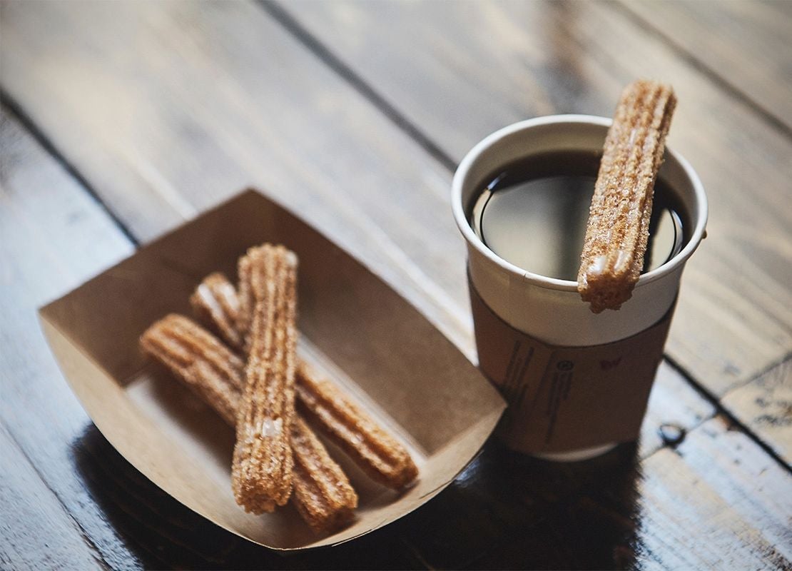 Vaso de café caliente acompañado de churros en una mesa de madera