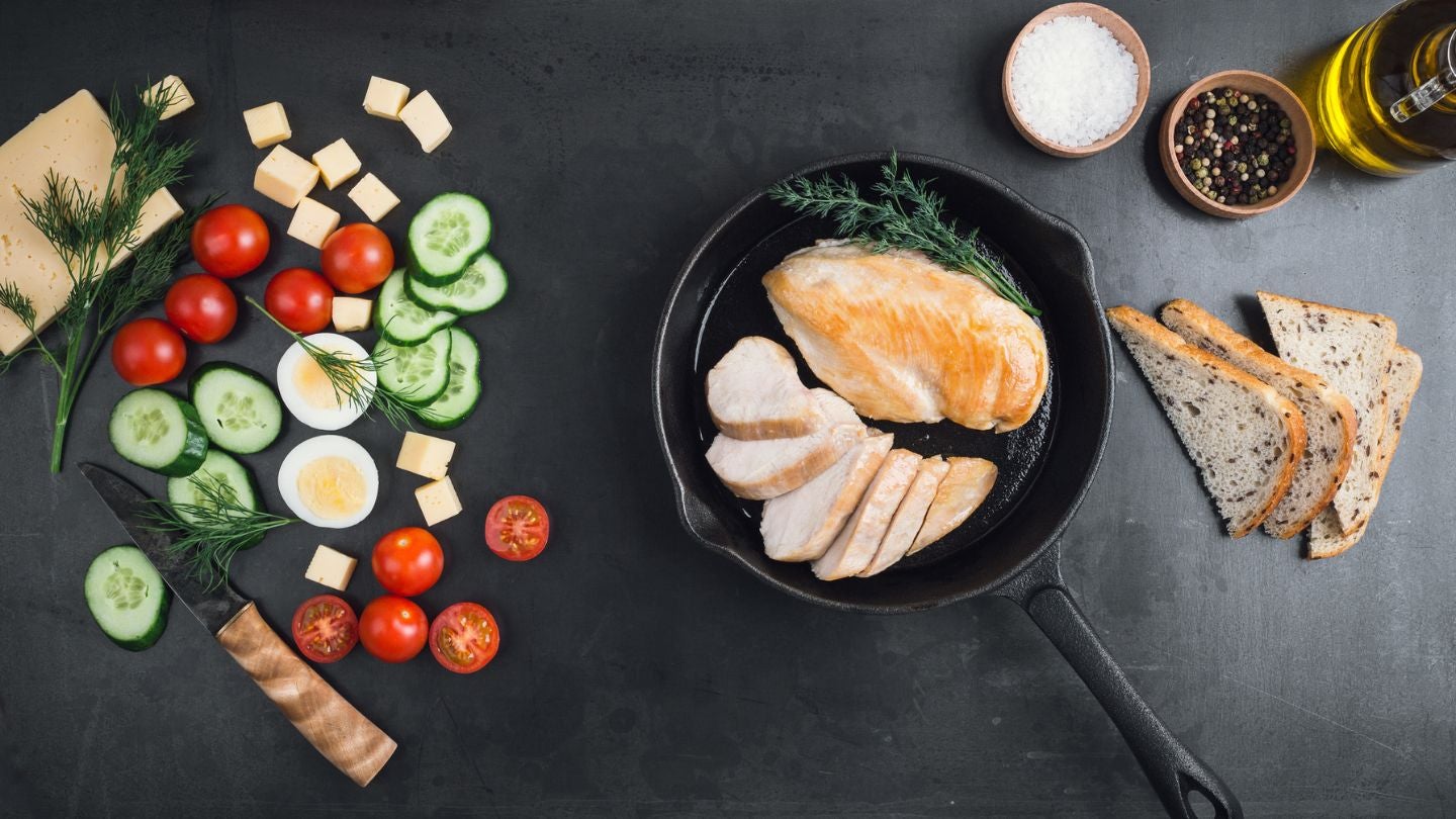 Sartén con pollo rodeado de verduras frescas, pan y especias sobre una mesa negra