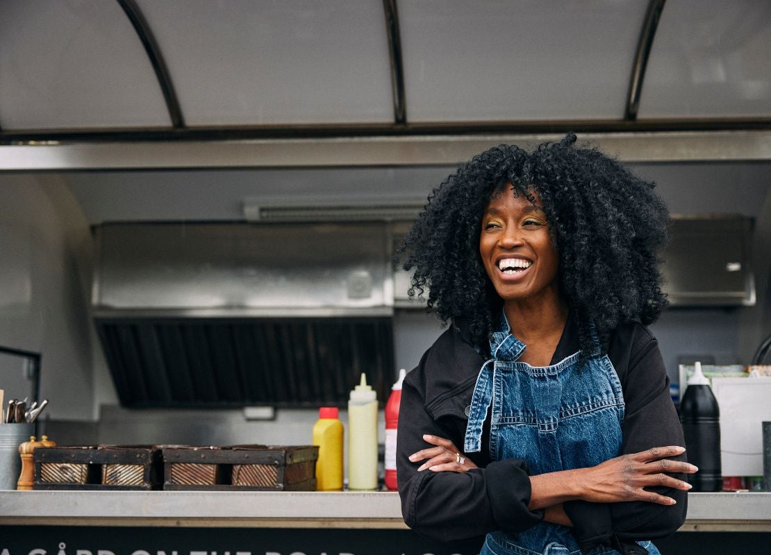 Persona sonriente frente a un food truck con insumos y estación de trabajo al aire libre.
