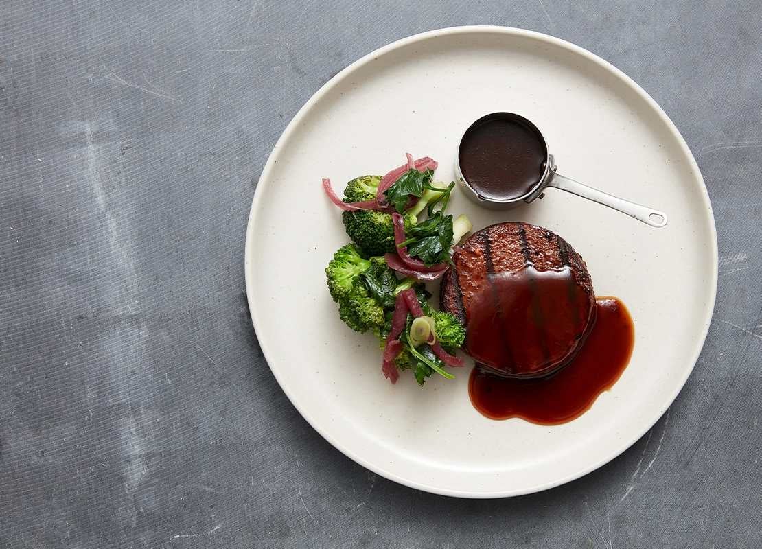 Hamburguesa sin pan bañada en salsa, servida con brócoli, cebolla encurtida y un recipiente con más salsa