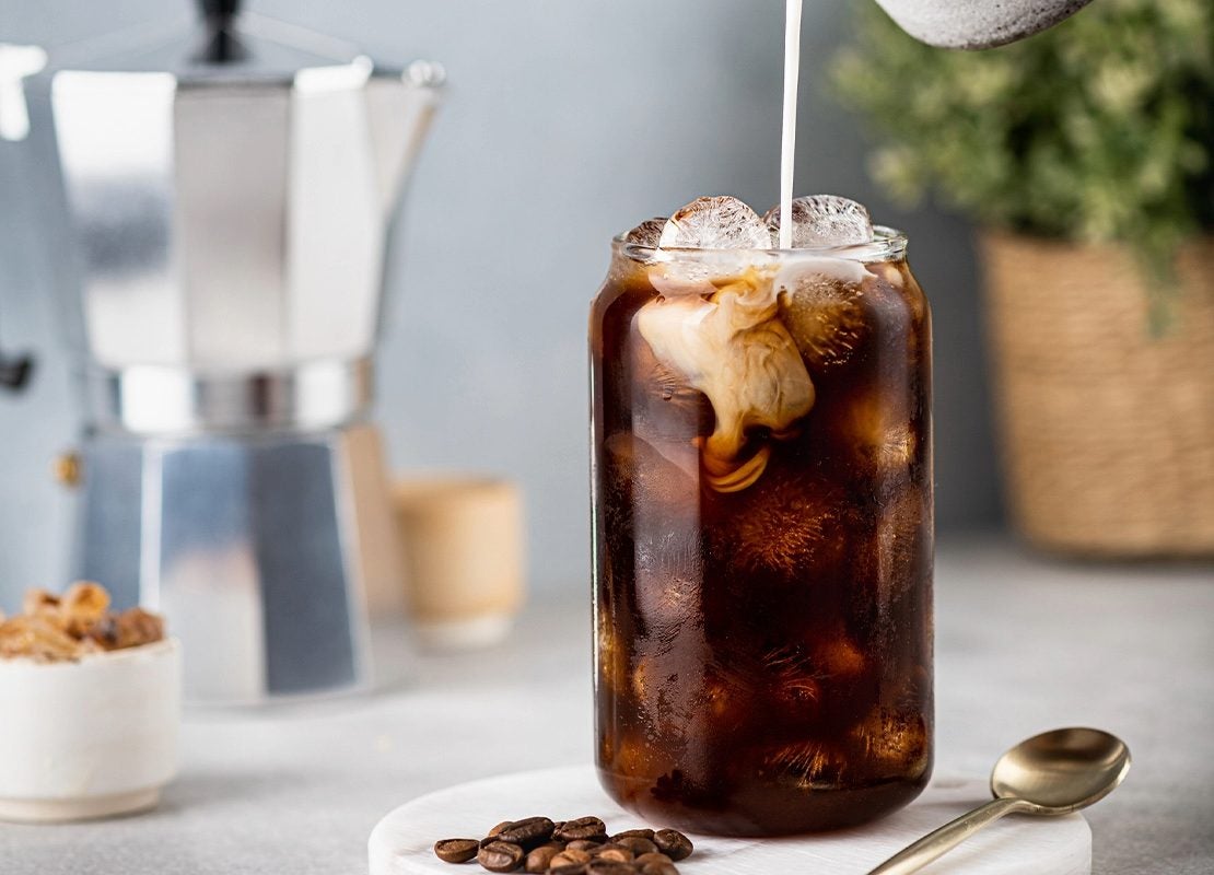 Persona vertiendo leche en un vaso con café frío y hielo, junto a una cafetera, granos de café y plantas