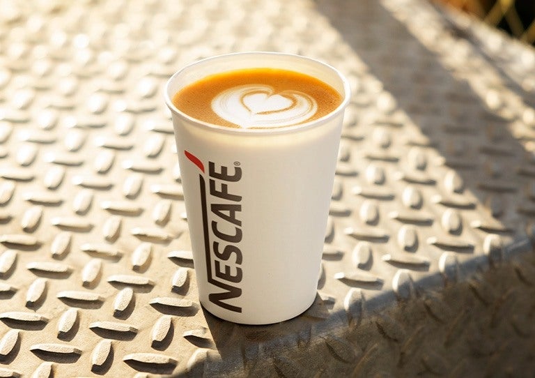Vaso de café con latte art y logotipo NESCAFÉ® sobre superficie metálica al aire libre