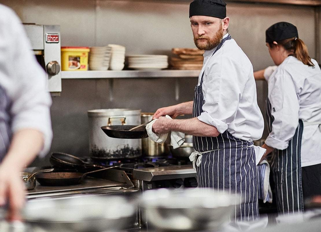 Chef ejecuta preparaciones en una estación caliente de una cocina industrial, rodeado de otros chefs