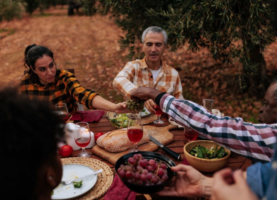 Personas compartiendo comida en mesa al aire libre con pan, uvas y bebidas de la dieta mediterránea