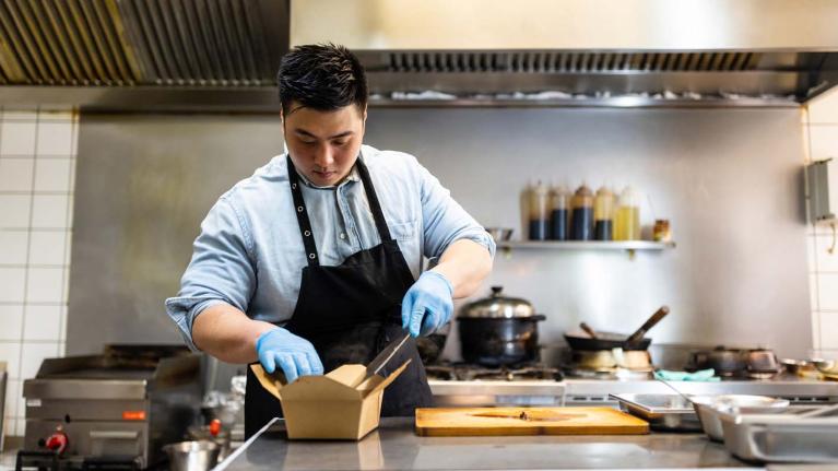 En una cocina comercial, chef empaca una preparación en una caja para ser enviada como entrega a domicilio