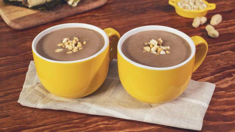 Dos tazas amarillas de chocolate caliente coronadas con cacahuates, al fondo hallacas sobre una tabla
