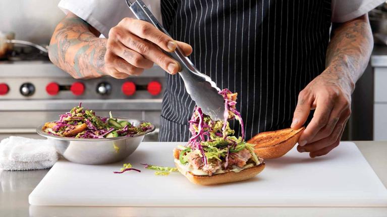 En una cocina comercial, chef agrega ensalada con pinzas al pan sobre una tabla de cortar