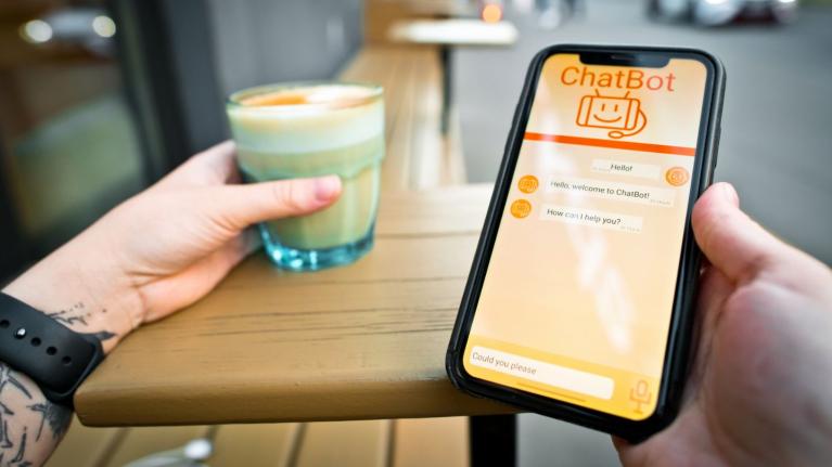 Persona sostiene celular con un chatbot en pantalla mientras toma una bebida en una cafetería