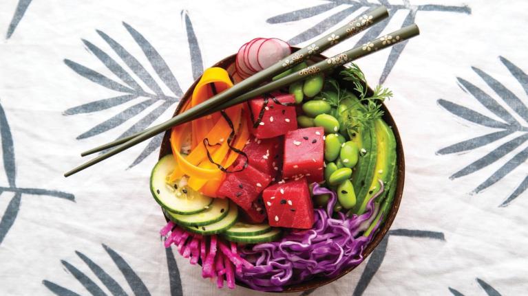 Bowl colorido de vegetales frescos y cubos de atún con palillos sobre un mantel decorado.