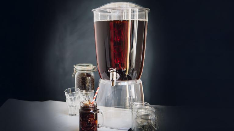 Dispensador de café cold brew junto a vasos y frascos con granos de café en una mesa.