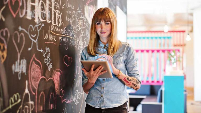 Mujer usa una tablet junto a un tablero con ideas en un espacio creativo.