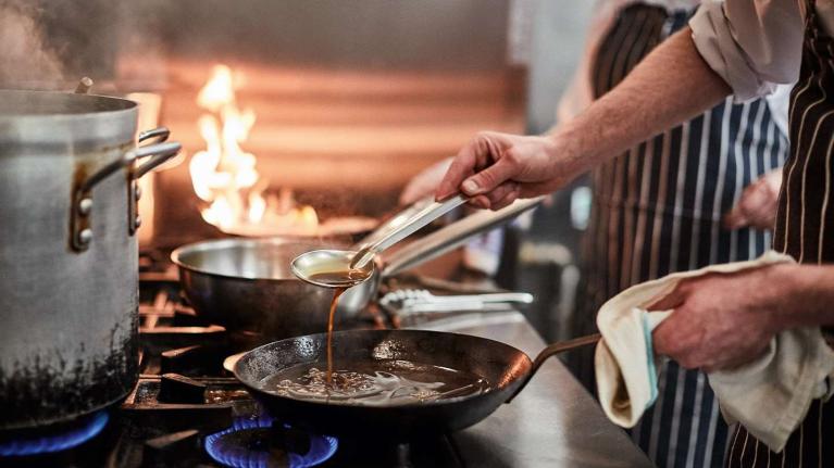 Chef vertiendo salsa en sartén caliente dentro de una cocina profesional en plena preparación