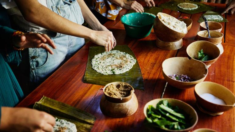 Personas preparando talames maya sobre hojas con ingredientes en recipientes sobre mesa de madera