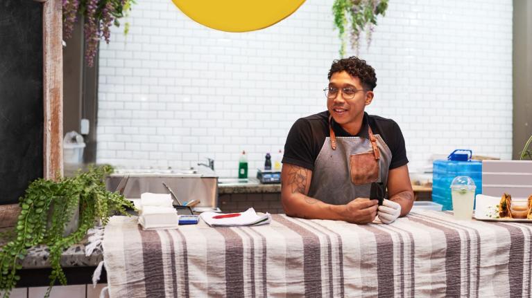 Hombre con delantal apoyado en mostrador de cocina con utensilios y plantas