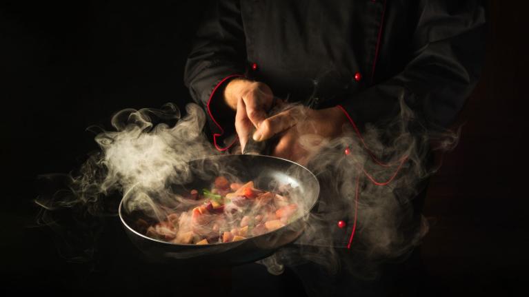 Chef sostiene una sartén emanando nubes de humo y vapor sobre ingredientes picados.