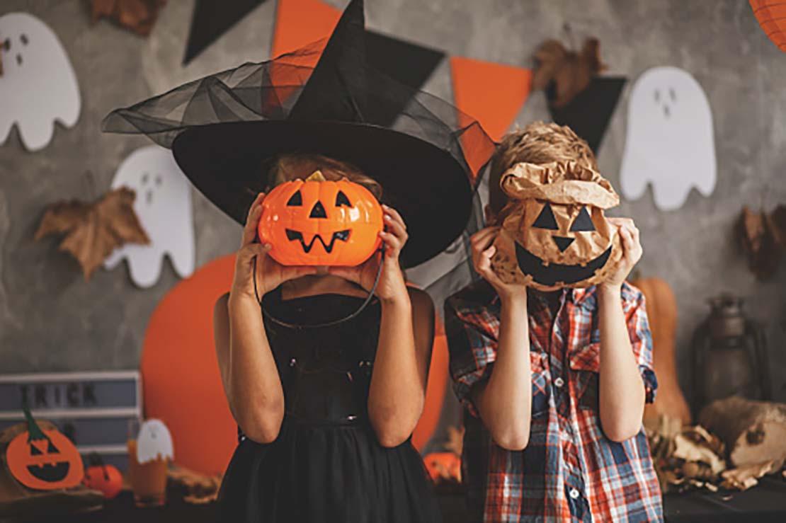 Dos niños que están en un espacio decorado para una fiesta de Halloween ocultan su rosto tras una calabaza y una bolsa