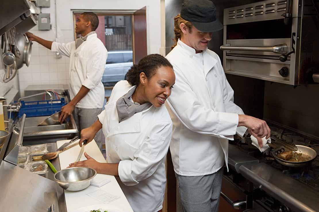 En una cocina industrial, una chef observa cómo otro fríe una proteína mientras un chef más lava las ollas