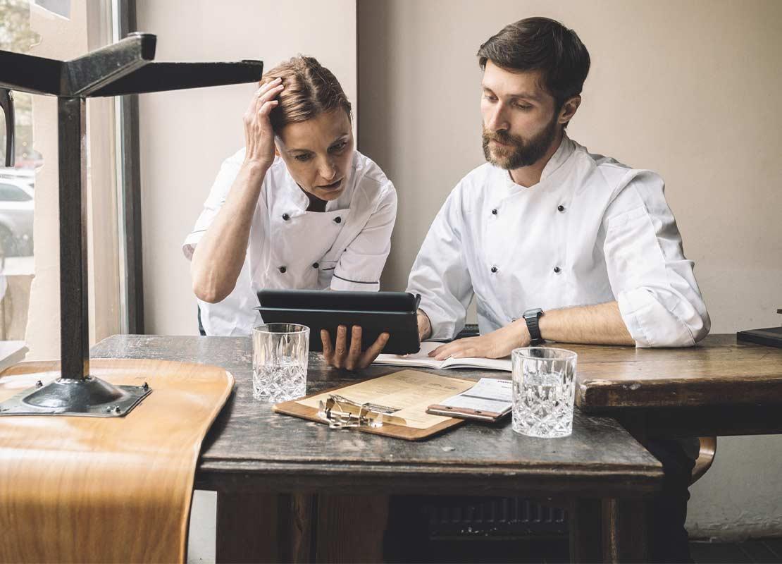 Dos chefs ejecutivos sentados a la mesa de un restaurante revisan en una Tablet el menú, platillos y presupuesto