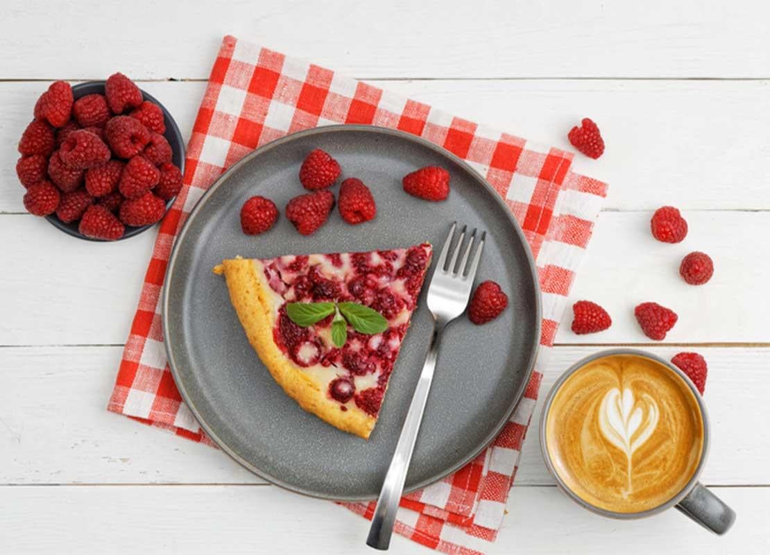Trozo de tarta de frambuesa, taza de café y cuenco de frambuesas sobre mesa de madera blanca que hacen buen maridaje