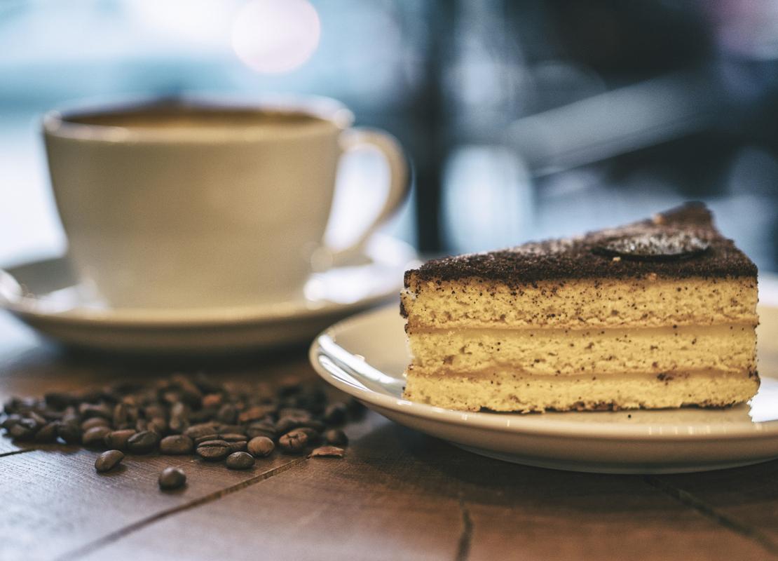 Una taza de espresso y, al frente, un trozo de tarta en una mesa de madera sobre la que hay regados granos de café tostados