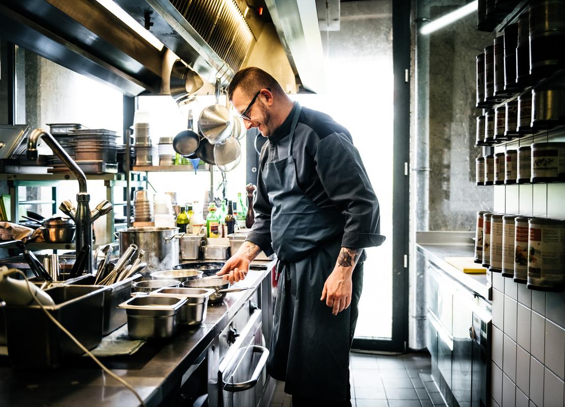 Chef revisa ollas que están sobre la estufa de una cocina de restaurante y frente a él hay utensilios en repisas y ganchos