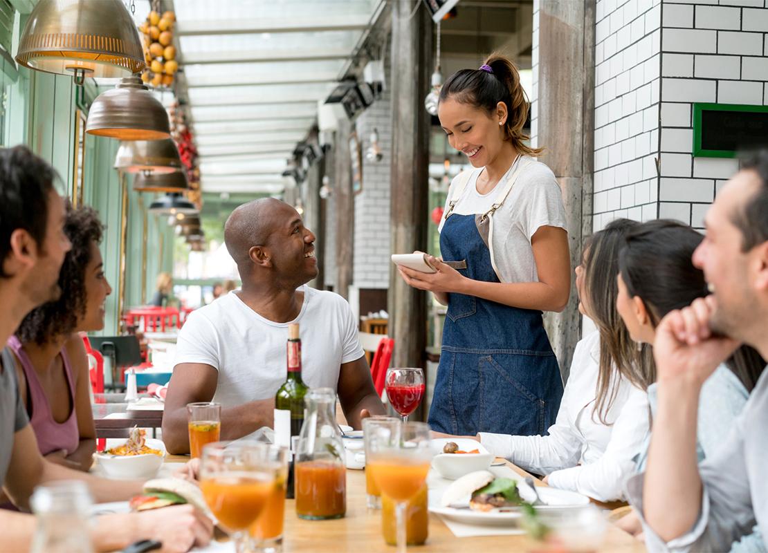 Mesera sonriente toma la orden de una mesa con 6 personas que la observan con emoción en un restaurante