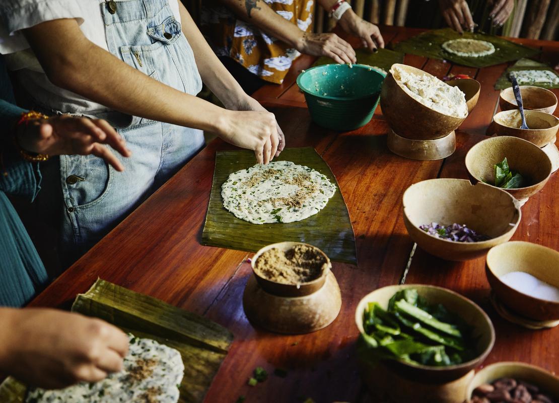 Brazos de personas preparan tamales oaxaqueños en una mesa usando hojas de plátano, masa de maíz y cuencos tradicionales