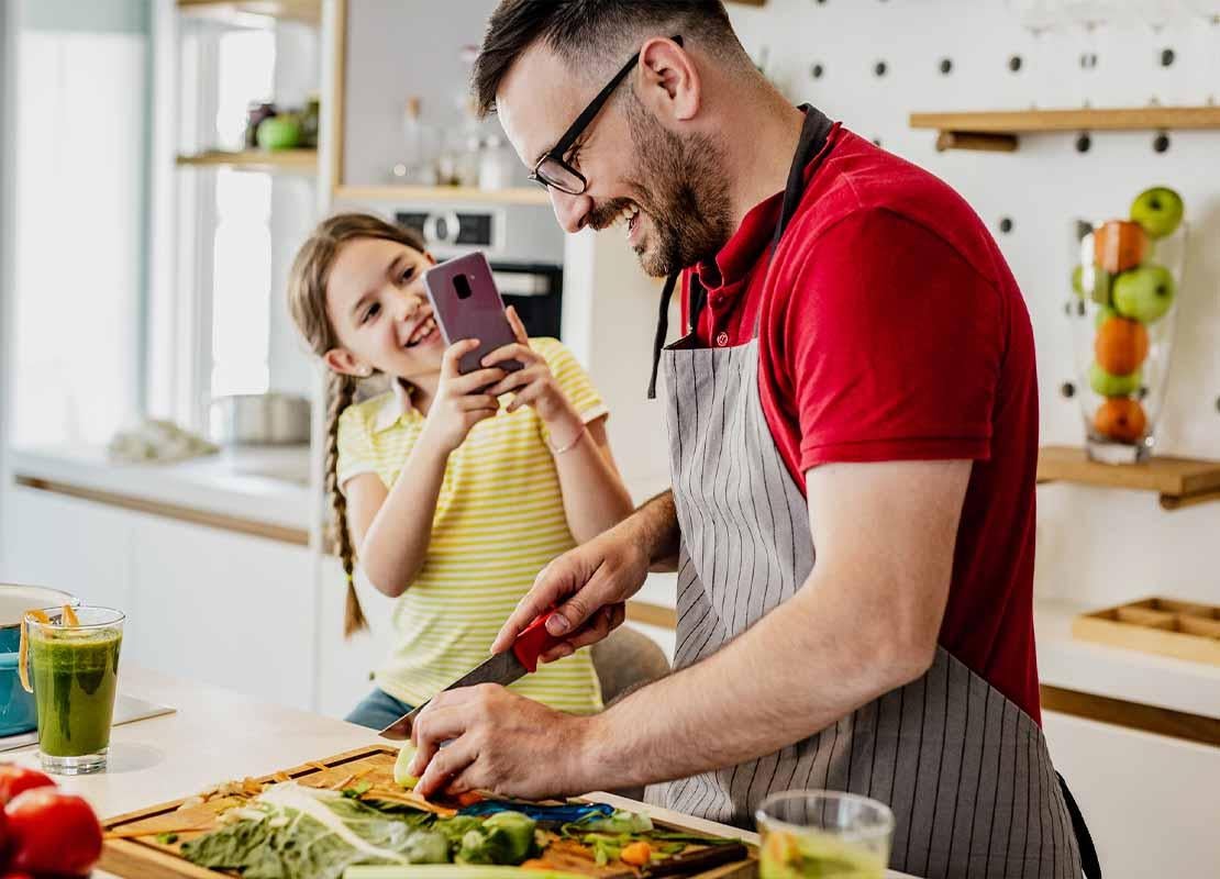 Hombre corta vegetales mientras su hija lo fotografía con un smartphone en un taller de cocina del Día del Padre